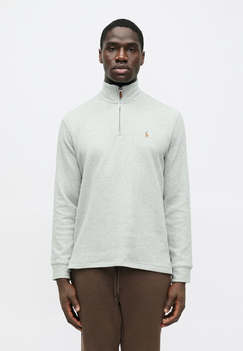 Hellgrauer Reißverschluss-Pullover aus weichem Stoff, mit einem hohen Kragen und einem kleinen gestickten Logo auf der Brust. Lange Ärmel.