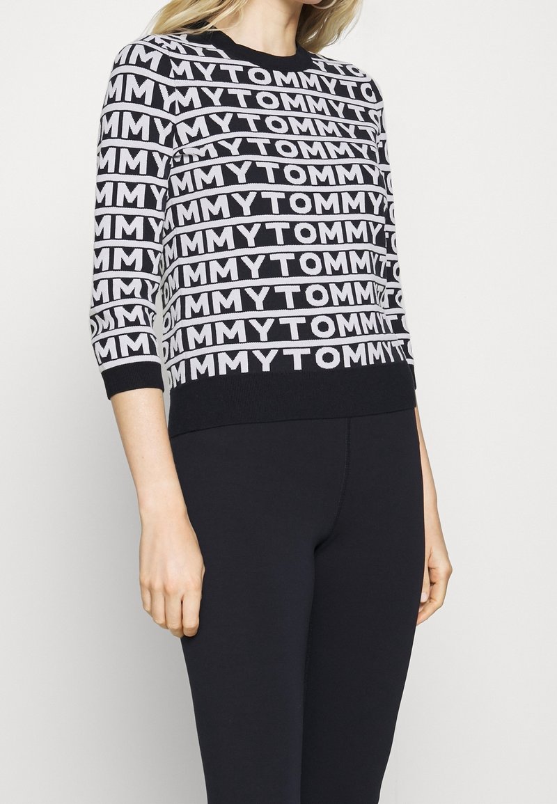 Schwarzer Pullover mit weißem, wiederholtem "TOMMY"-Text, runder Ausschnitt und Dreiviertelärmeln. Weiches Strickmaterial, schlichte Gestaltung.