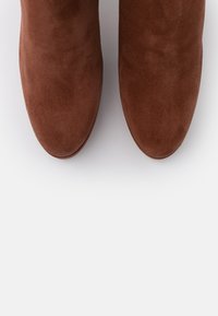 Botas de tobillo de ante marrón con una textura suave, punta redondeada y diseño minimalista. Sin herrajes ni patrones visibles.