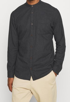 Mężczyzna ubrany w ciemnoszarą, zapinaną na guziki koszulę z długim rękawem, kołnierzem typu mandarynka oraz kieszenią na piersi, w połączeniu z beżowymi spodniami.