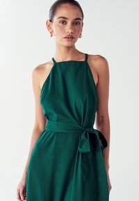 Zelena halter haljina od teksturiranog materijala, s uklopljenim korzetom, pojasom na struku i otvorenim dizajnom na leđima. Zlatne kružne naušnice kao dodatak.