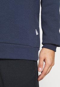 Felpa blu navy con orlo e polsini a coste. Etichetta visibile sul lato inferiore. Tessuto liscio con design aderente, indossata con pantaloni scuri a motivo.
