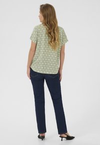 Blusa verde con estampado y mangas cortas, combinada con unos jeans de mezclilla azul oscuro. La blusa presenta un diseño geométrico y un corte holgado.