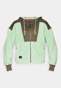 Fleece hettejakke med glidelås i lysegrønn og mørk brun med paneler, snorhette og strukturert overflate. Svart logo-lapp på kanten.