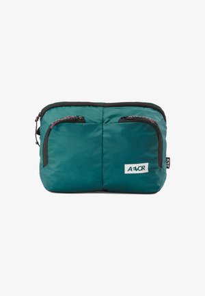 Sac ceinture compact bleu sarcelle avec deux poches avant zippées et une bordure noire, arborant un patch logo AEVOR blanc en bas à droite.
