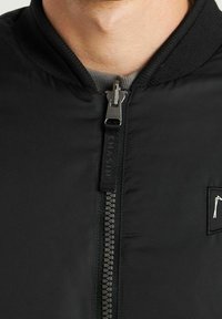 Close-up van een persoon in een zwarte ritsjack met geribbelde kraag en een klein rechthoekig patch op de borst.