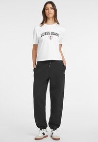 Witte T-shirt met korte mouwen met de tekst "GUESS JEANS", gecombineerd met zwarte sweatpants met elastische boorden en witte sneakers met zwarte en bruine accenten.