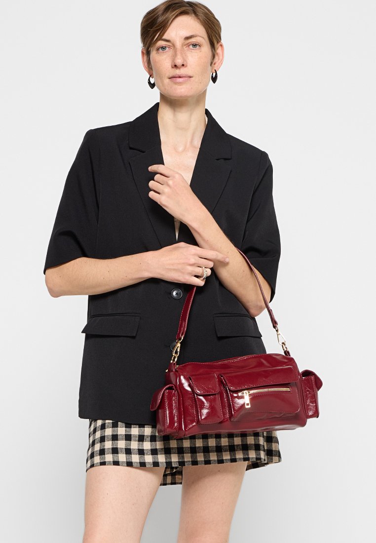 Burgunderfarbene Lacklederhandtasche mit goldenen Beschlägen, Reißverschlussfach und zwei seitlichen Taschen. Kompakte rechteckige Form, strukturiertes Design.
