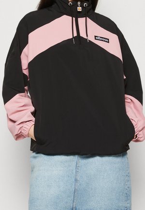 Persoon draagt een zwarte en roze ellesse pullover jas met een ritssluiting aan de voorkant en lichtblauwe spijkerbroek, handen in de zakken.