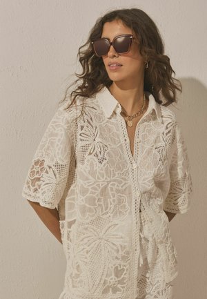 Kobieta w dużych brązowych okularach przeciwsłonecznych, złotym naszyjniku z zawieszką i białej koronkowej koszuli z krótkim rękawem zapinanej na guziki na tle beżowej ściany.