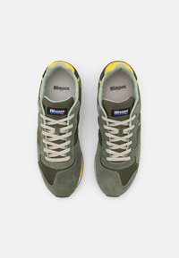 Baskets vert olive avec une tige en daim et mesh, lacets beige, accents jaunes et un talon noir, dotées d'une semelle intérieure de marque.