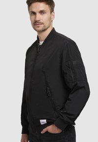 Schwarze Bomberjacke aus glattem, leichtem Stoff mit gerippten Bündchen, Reißverschluss vorne und Seitentaschen. Mit Markenzeichen am Ärmel.