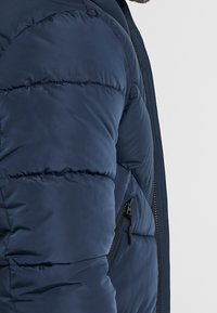 Marineblauwe pufferjack met gewatteerde textuur en horizontale stiksels. Voorzien van een verticale zak met rits aan de zijkant en een gladde, waterafstotende afwerking.