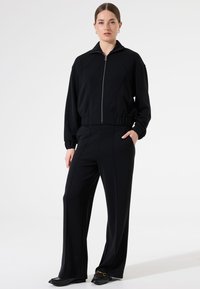 Schwarzes Sportset mit einer Reißverschlussjacke mit Kragen und elastischem Bund, kombiniert mit weit geschnittenen schwarzen Hosen und flachen Schuhen.