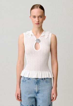 Giovane donna con i capelli raccolti in uno chignon indossa un top in maglia bianco senza maniche con apertura a goccia e spilla floreale, abbinato a jeans denim azzurri chiari.