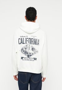 Balta džemperis su tekstūruotais grafika ant nugaros, kuriame užrašyta "Welcome To CALIFORNIA", atvaizduotas lokys, o apačioje - "Tommy Jeans".