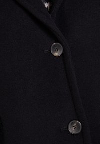 Wolblend jas in donker marineblauw met een gestructureerd oppervlak. Kenmerkt zich door twee zwarte knopen, gekraagde schouders en zijzakken. Klassiek op maat gemaakt ontwerp.