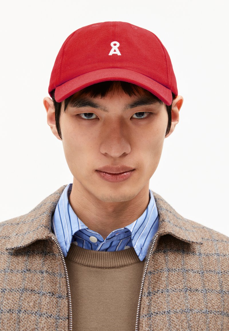 Casquette de baseball en coton rouge avec une visière courbée et un logo brodé blanc. Portée avec une veste à carreaux marron sur une chemise rayée bleue.