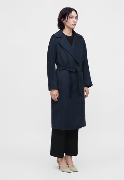 Femme debout portant un trench-coat bleu marine foncé ceinturé, un pantalon noir à jambes larges et des escarpins pointus beige clair sur un fond uni.