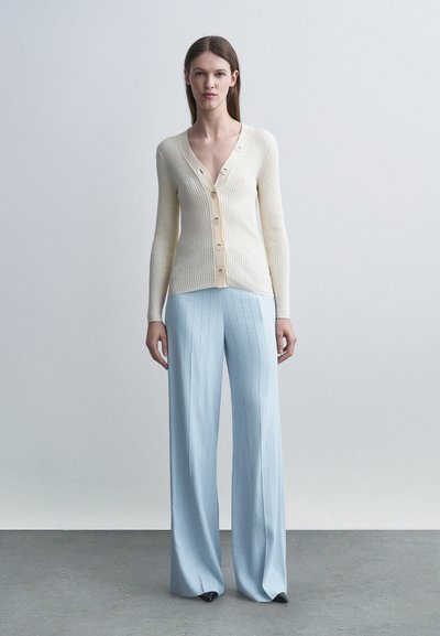 Γυναίκα όρθια φοράει εκρού ζακέτα με V λαιμόκοψη και ρίγες, με κουμπιά, και ανοιχτό μπλε παντελόνα σε απλό γκρι φόντο.