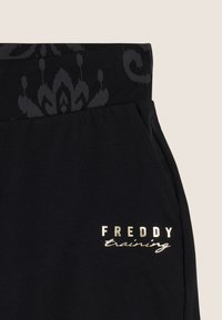 Pantalones deportivos negros con una cinturilla estampada y un logo metálico plateado "FREDDY training" en el bolsillo delantero. Material de algodón suave.