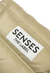 Beige gepolsterter Stoff mit einem schwarz-weißen Etikett, auf dem "SENSES .THE LABEL" steht, sichtbaren Nähten und einer glatten Textur.