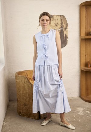 Žena nosi svetlomodré pruhované šaty bez rukávov so zviazanými mašľami, stojaca vo vnútri blízko drevenej stoličky a knižnice.