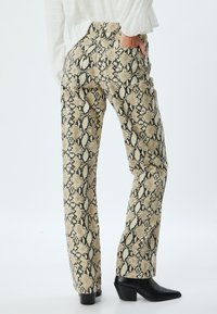 Pantalons beiges avec un motif imprimé serpent noir, coupe droite et trois poches. Portés avec des bottines à talons noires.