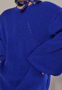 Pull en maille bleu surdimensionné avec texture côtelée, présentant des motifs en diamant et de petites découpes. Manches larges et coupe décontractée.