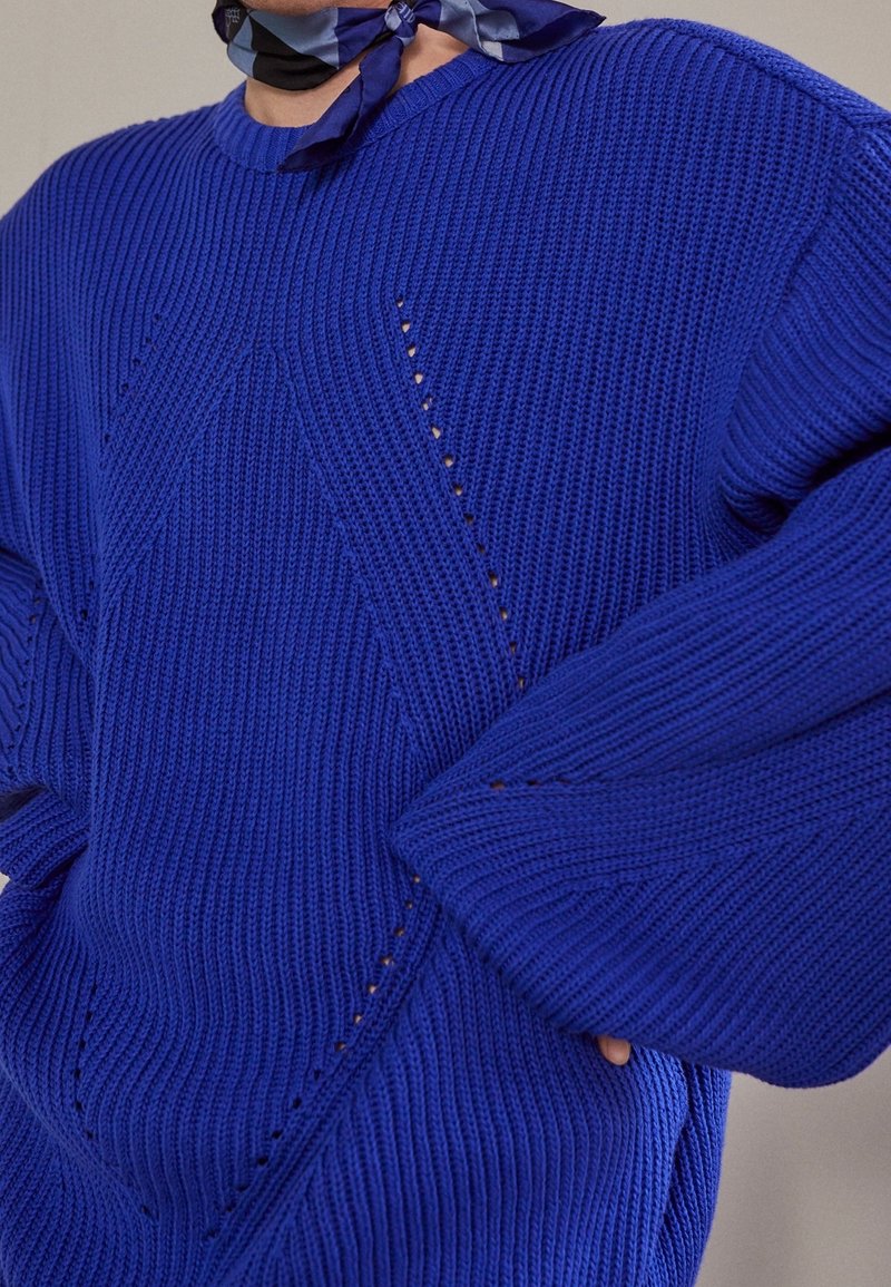 Pull en maille bleu surdimensionné avec texture côtelée, présentant des motifs en diamant et de petites découpes. Manches larges et coupe décontractée.