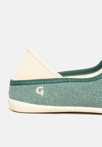 Green canvas slip-on shoe with a light beige accent at the heel, featuring a textured surface and a small embroidered "G" logo.