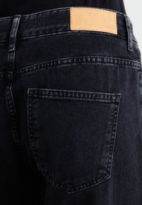 Pogled od blizu na zadnji žep črnih denim hlač z vidnimi šivi in svetlo rjavim usnjenim blagovnim znakom na pasu.