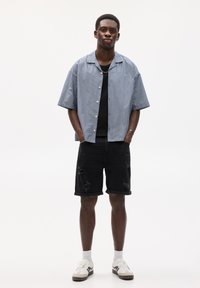 Chemise à manches courtes bleu et blanc rayée, débardeur noir, shorts noirs usés, baskets blanches avec des accents gris, se tenant sur un fond neutre.
