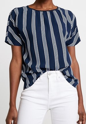 Chemise bleu marine à manches courtes avec des rayures blanches verticales, portée rentrée dans un pantalon blanc taille haute sur une personne.