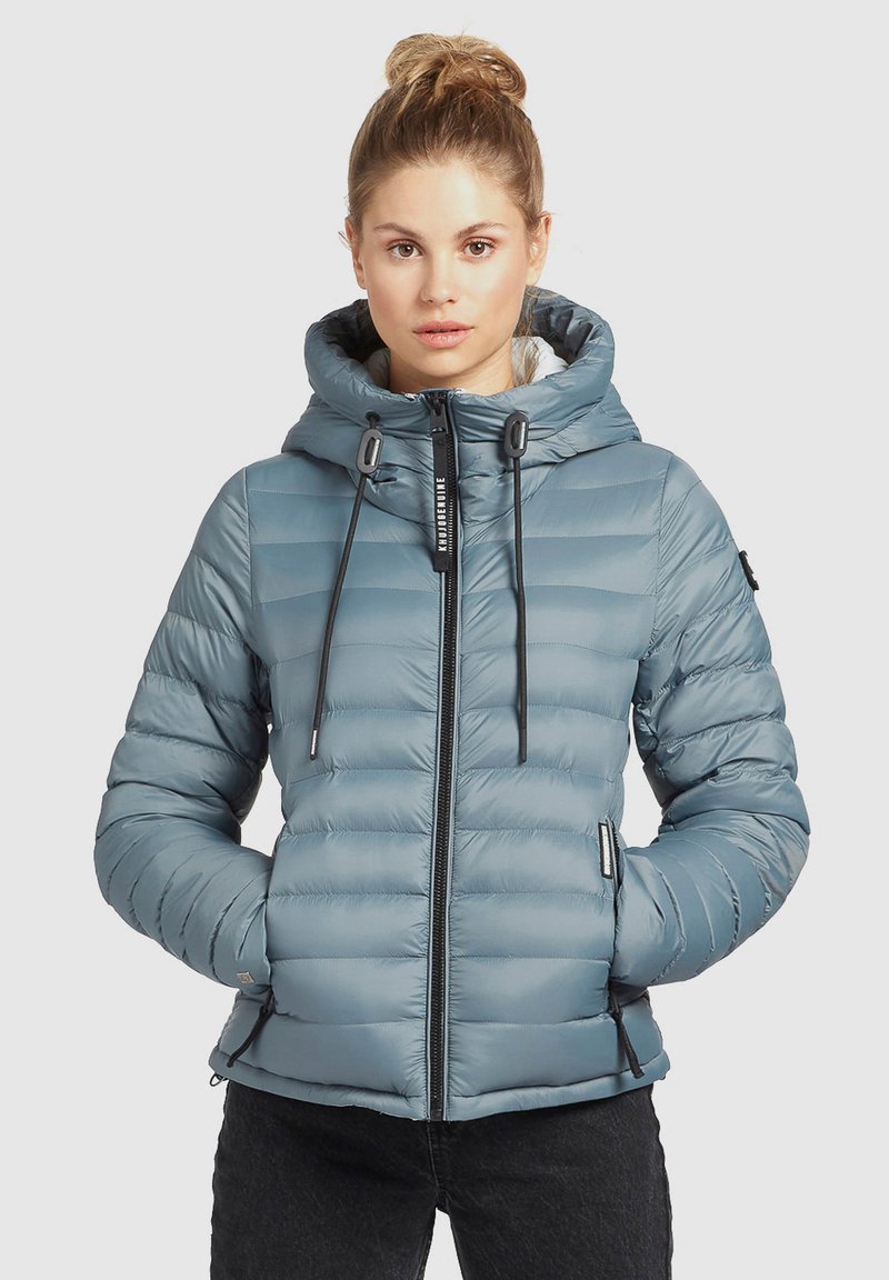 Helles blauer Pufferjacke mit Kapuze, ausgestattet mit einem Reißverschluss vorne, Kordelzügen und Seitentaschen. Horizontale Steppstruktur im gesamten Material.