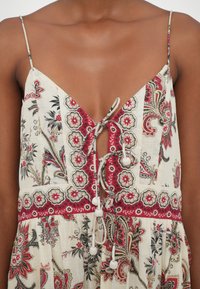 Femme portant une robe sans manches avec des motifs floraux et cachemire en rouge, noir et blanc, avec un détail noué à l’avant orné de pompons.
