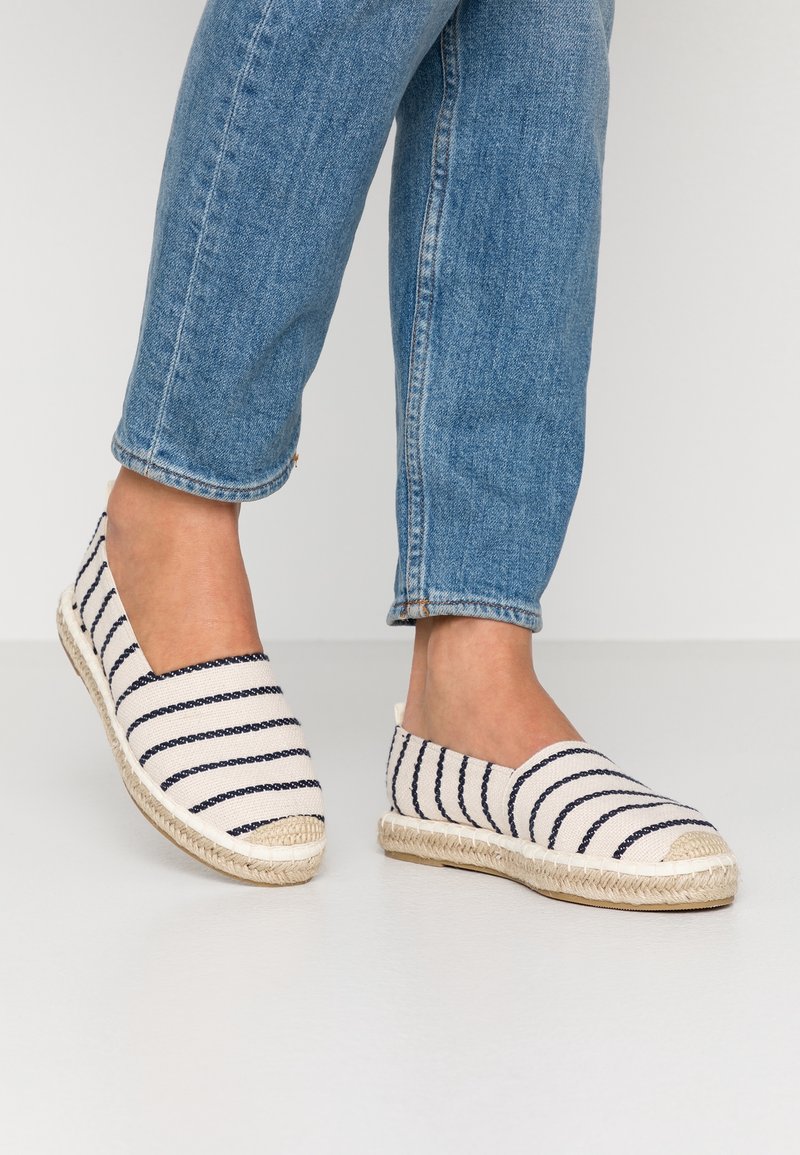 Canvas espadriller med marinblå och vita ränder, rund tå och sula av jute-rep. Bar med uppvikt denimjeans, vilket visar avslappnad fotposition.