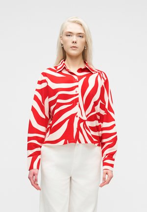 Kvinde med langt blondt hår iført en rød og hvid zebra-stribet skjorte med knapper og hvide bukser, stående foran en ensfarvet baggrund.