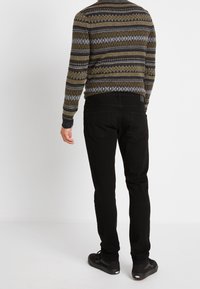 Jeans negros y un suéter con un diseño estampado que presenta zigzags y diamantes en oliva, azul y gris, con puños acanalados y un cuello redondo.