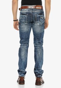 Jean en denim bleu pour homme, coupe slim, délavé, cinq poches, ceinture en cuir marron, coutures visibles et léger délavage près des genoux.