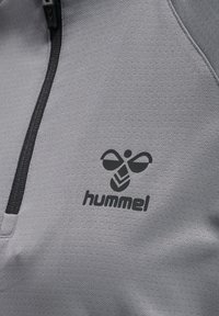 Pullover sportivo grigio con mezzo zip, caratterizzato da un tessuto testurizzato e un logo nero di un'ape stilizzata con la scritta "hummel."