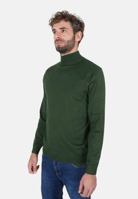 Maglione a collo alto verde realizzato in tessuto morbido, con maniche lunghe e orlo a coste. Abbinato a jeans di denim blu.
