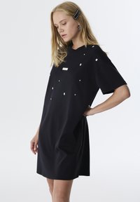 Schwarzes kurzärmeliges Kleid in lässiger Passform, verziert mit verstreuten Strasssteinen und einem rechteckigen Logo-Tag auf der Vorderseite für einen strukturierten Look.