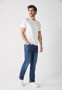 Camisa henley blanca de manga corta con una tapeta de tres botones y un pequeño logo amarillo, combinada con jeans azul oscuro y zapatillas blancas.