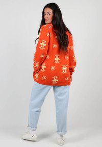 Un pull en tricot orange avec un motif beige répétitif de soleils et d'yeux, associé à un jean baggy bleu clair et des baskets blanches.