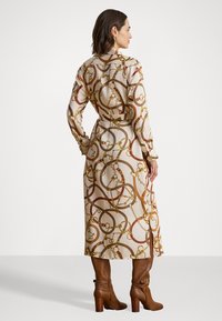 Abito a maniche lunghe in tessuto beige con un motivo a catena circolare in oro, rosso e marrone, abbinato a stivali alti fino al ginocchio con tacco marroni.