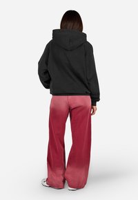 Čierna mikina s uvoľneným strihom a nastaviteľnou kapucňou, skombinovaná so širokými nohavicami v prechode červenej a ružovej farby. Mäkký, textúrovaný materiál.