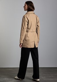 Valkoinen trench-coat, jossa on vyöllinen vyötärö, kaulus ja pitkät hihat, yhdistettynä mustiin leveisiin housuihin ja valkoisiin Tennareihin.