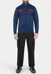 Veste zippée bleu marine avec rayures rouges, poignets et col élastiques ; associée à un pantalon cargo noir avec poches ; portée avec des baskets foncées.