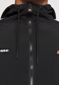 Gros plan d'une veste à capuche noire avec une fermeture éclair centrale et des cordons noirs, présentant un patch logo blanc sur le côté gauche.